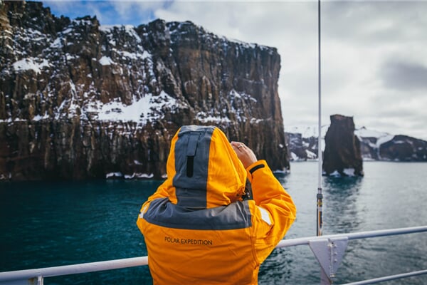 Guest in yellow parka stands on the main deck of their ship and takes a photograph with their smartphone. Sea days offer the best opportunities for photography of sea birds, icebergs and rock formations!